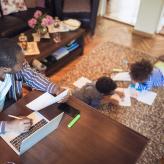 Adult man works on laptop and looks after two children reading nearby in room.