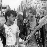 Two people at a protest. One is holding a flag, wearing a leather jacket. The other man is wearing a white t-shirt with anti-nazi writing. 